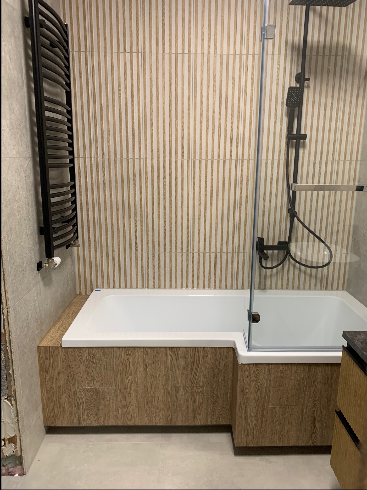 Nowoczesna łazienka po remoncie: biała wanna z drewnopodobną obudową, czarna armatura i grzejnik na tle płytek w pionowe pasy. Minimalistyczny design.