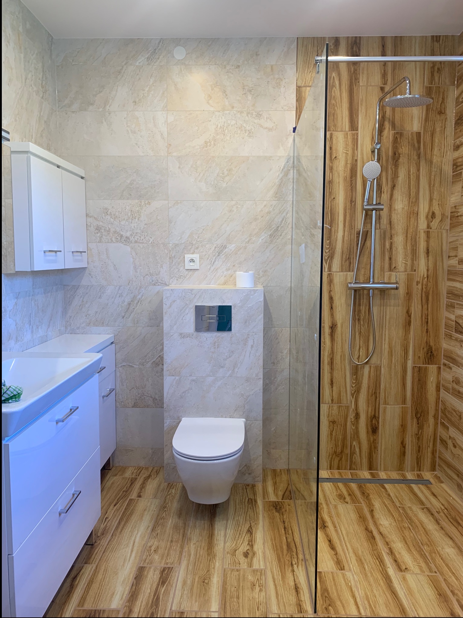 Nowoczesna łazienka z kabiną prysznicową typu walk-in, wykończona beżowymi i drewnopodobnymi płytkami, biała umywalka z szafką i podwieszana toaleta.