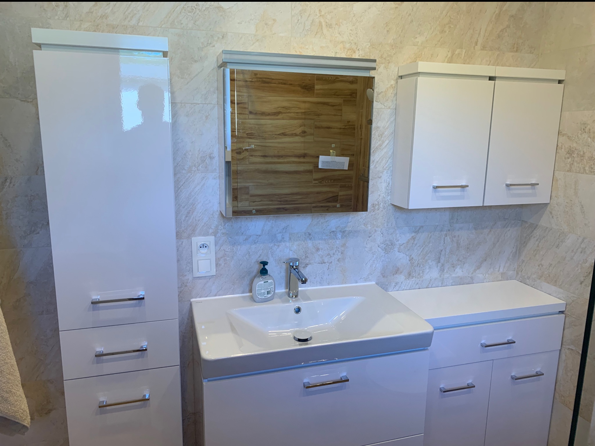 Białe szafki łazienkowe z umywalką i lustrem na tle beżowych płytek. Nowoczesny design, chromowane uchwyty i bateria. Widoczny dozownik mydła i gniazdko.