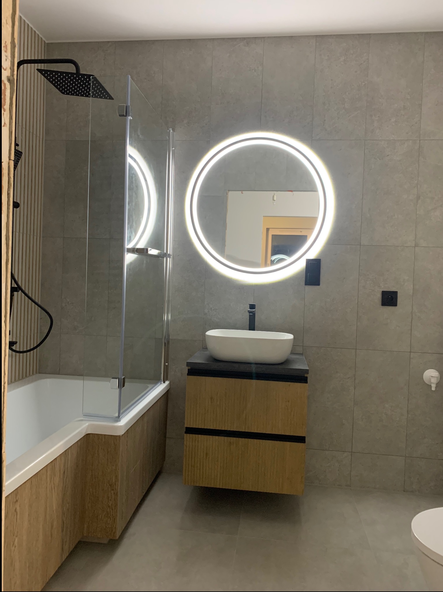 Nowoczesna łazienka: szare płytki, okrągłe lustro z LED, biała umywalka nablatowa na szafce z drewnopodobnym frontem, czarna armatura i kabina prysznicowa.