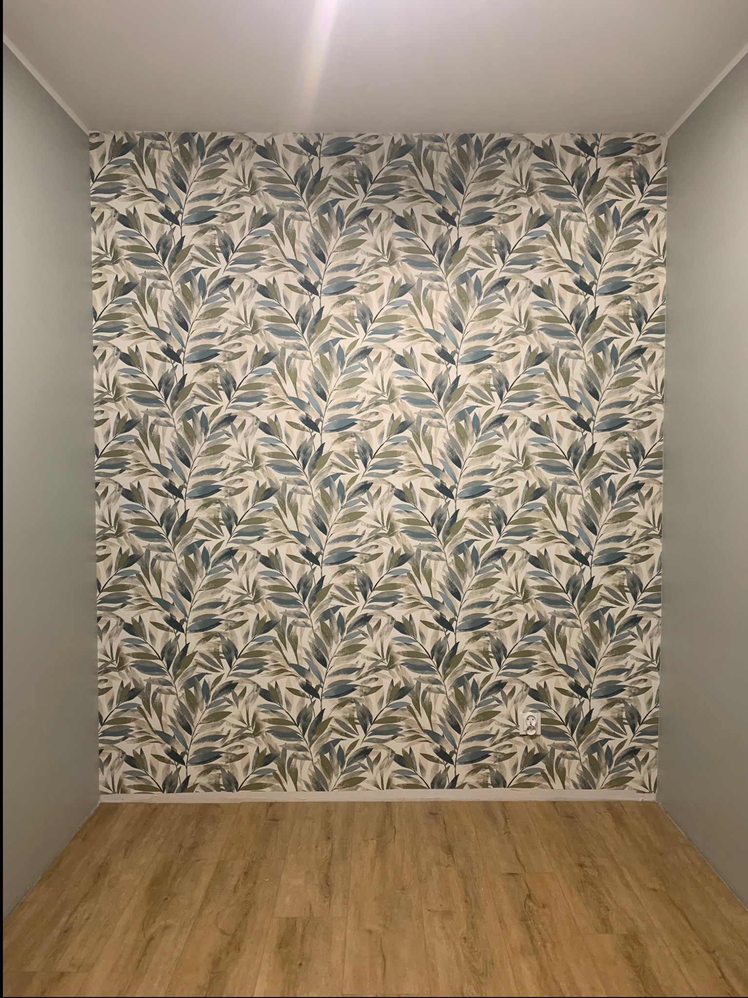 Pokój po remoncie: ściana w liście, drewniana podłoga, szare ściany boczne. Widoczny kontakt elektryczny. Jasne oświetlenie sufitowe.