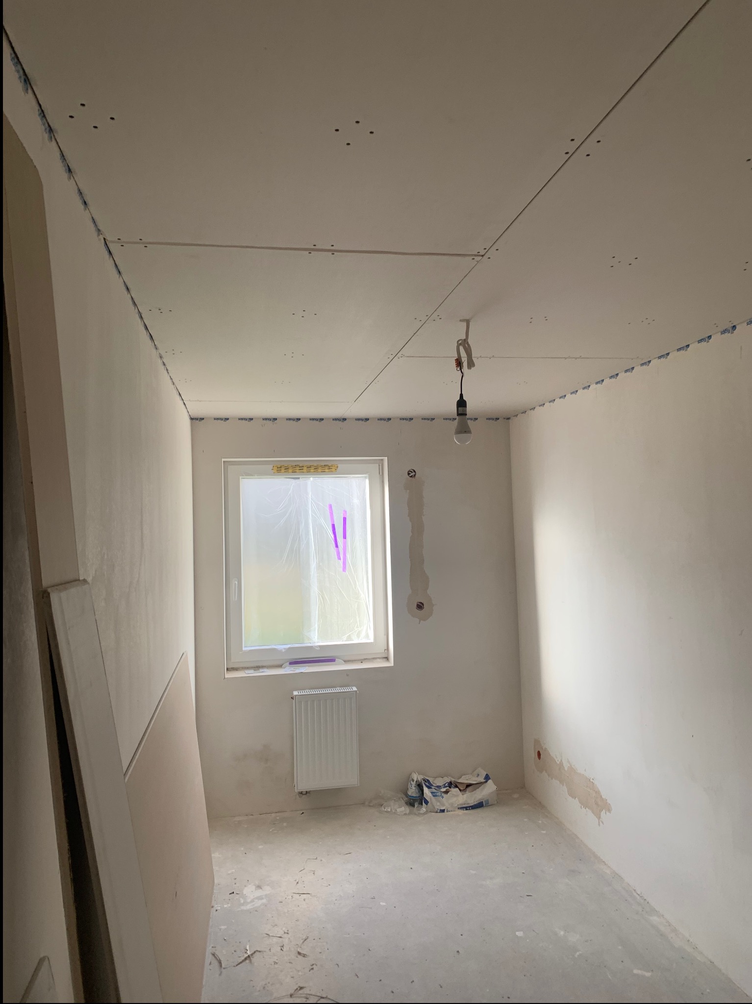 Pokój w trakcie prac wykończeniowych: zamontowane płyty gipsowo-kartonowe na suficie, ściany przygotowane do dalszych prac. Okno zakryte folią.