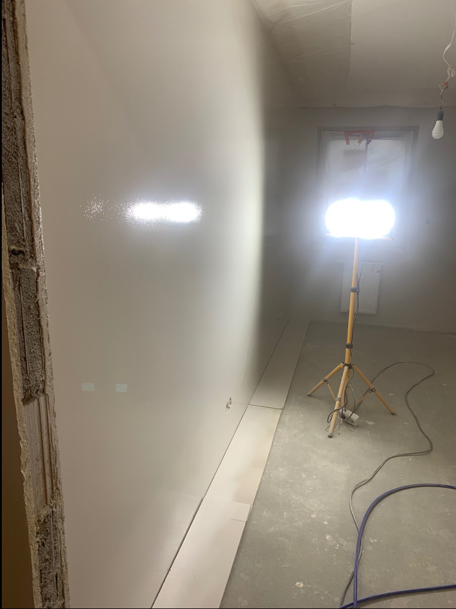 Świeżo wygładzona ściana w trakcie prac wykończeniowych, oświetlona lampą budowlaną. Widoczne ślady po szpachlowaniu i przygotowaniu do malowania.