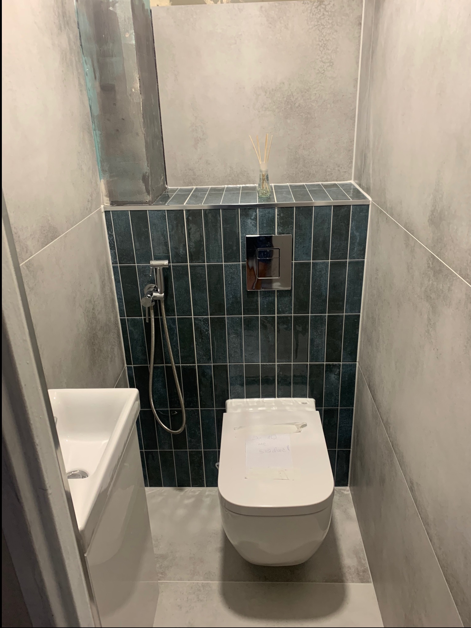 Nowoczesna toaleta z podwieszaną muszlą, zielona glazura w pionowy wzór, szara ściana z efektem betonu, minimalistyczna umywalka. Wykończona łazienka.