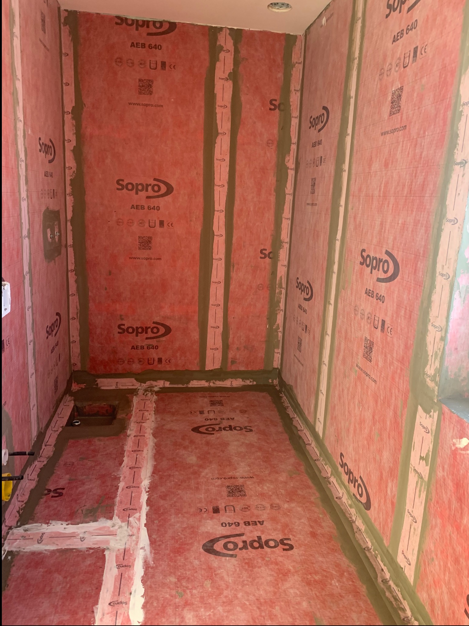 Przygotowanie do hydroizolacji w łazience: ściany i podłoga pokryte czerwoną membraną Sopra AEB 640, uszczelnione na łączeniach. Widok na wnętrze kabiny prysznicowej.