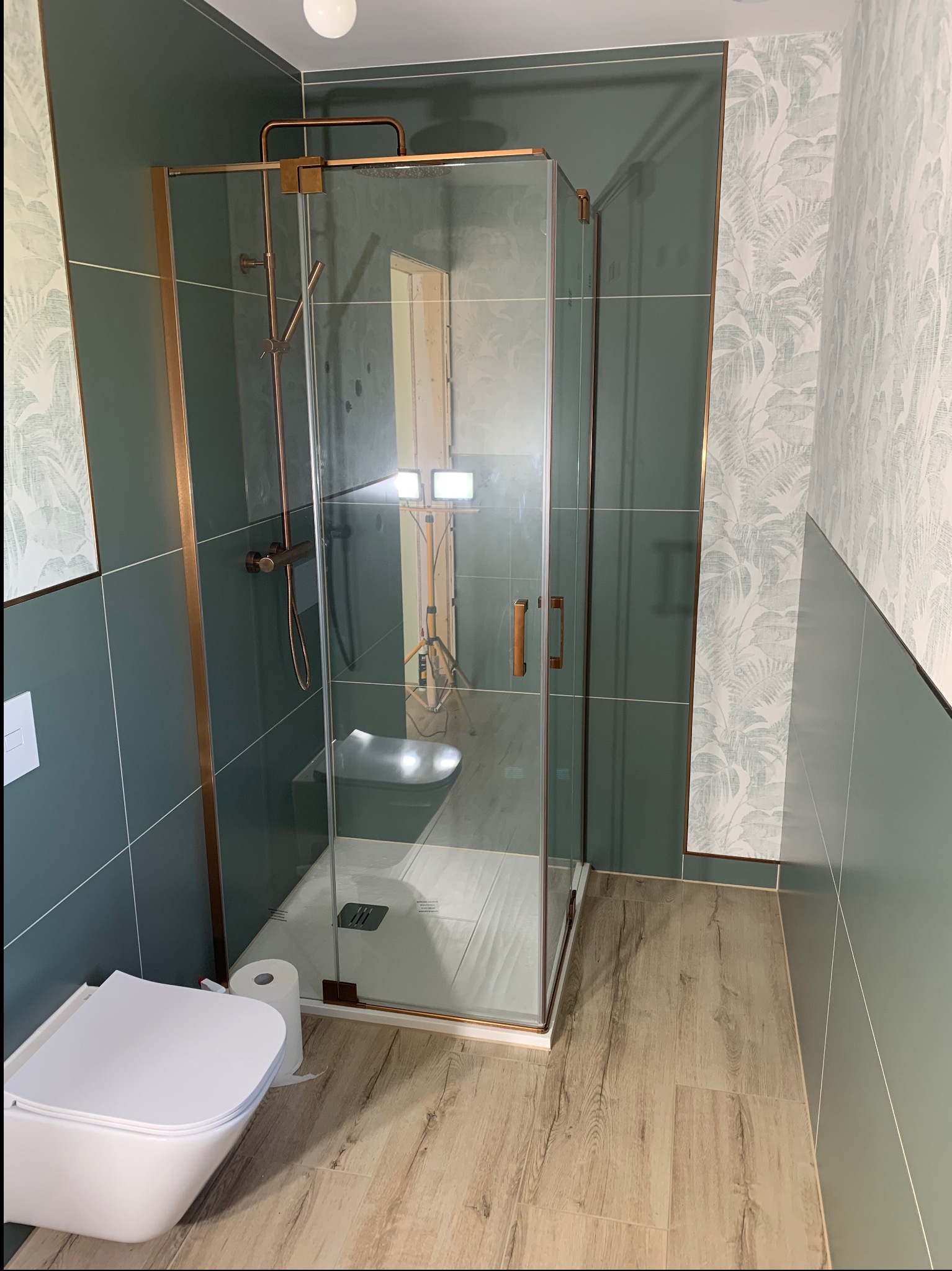 Wykończona łazienka z kabiną prysznicową z miedzianymi detalami, toaletą podwieszaną i panelami podłogowymi w odcieniach drewna. Ściany w dwóch kolorach, z motywem liści.