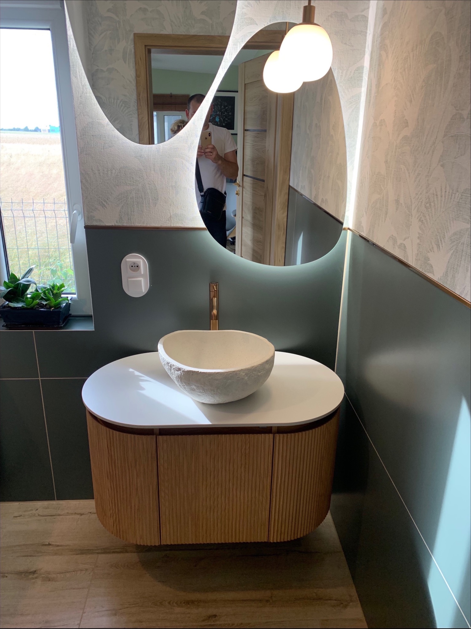 Nowoczesna łazienka z umywalką w kształcie misy, podświetlanym lustrem o nieregularnym kształcie i drewnianą szafką. Widoczne zielone akcenty i tapeta w roślinny wzór.