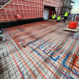 Karol Glica - Pomarańczowe rury instalacji podłogowej ułożone na siatce zbrojeniowej, widoczni pracownicy w odblaskowych ubraniach, budynek w tle.