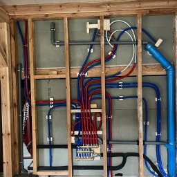 Karol Glica - Instalacja wodna w trakcie montażu: rozdzielacz, rury PEX w kolorach czerwonym i niebieskim, szare rury kanalizacyjne, widoczne elementy konstrukcji drewnianej ściany.