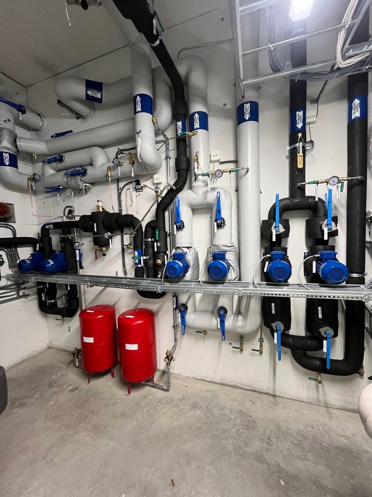 Kompleksowa instalacja grzewcza w kotłowni: rury w izolacji, zawory, pompy obiegowe, dwa czerwone zbiorniki wyrównawcze, manometry, zamontowane na ścianie w pomieszczeniu technicznym.