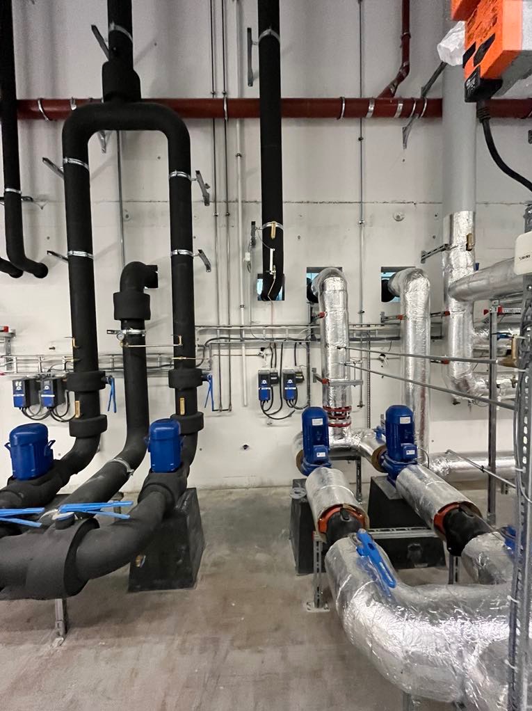 Szczegółowe ujęcie instalacji rurowej z izolacją termiczną, zawierającej czarne i srebrne rury, niebieskie zawory i liczne przewody, zamontowanej na jasnej, betonowej ścianie w pomieszczeniu...