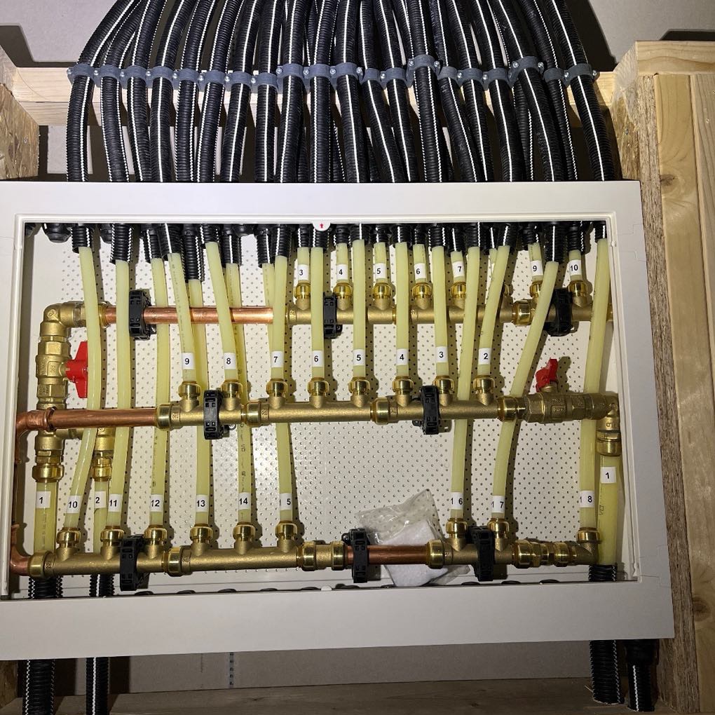 Rozdzielacz instalacji ogrzewania podłogowego z numerowanymi obwodami, widoczne rury doprowadzające i odprowadzające wodę, zamontowany w skrzynce z białym obramowaniem.