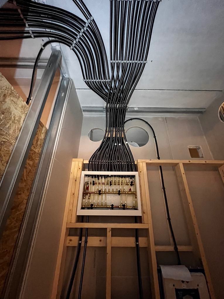 Rozdzielacz instalacji grzewczej podłączony licznymi czarnymi rurami, zamocowany w drewnianej konstrukcji w surowym wnętrzu podczas montażu.