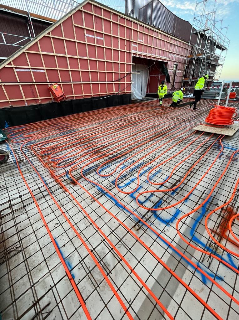 Pomarańczowe rury instalacji podłogowej ułożone na siatce zbrojeniowej, widoczni pracownicy w odblaskowych ubraniach, budynek w tle.