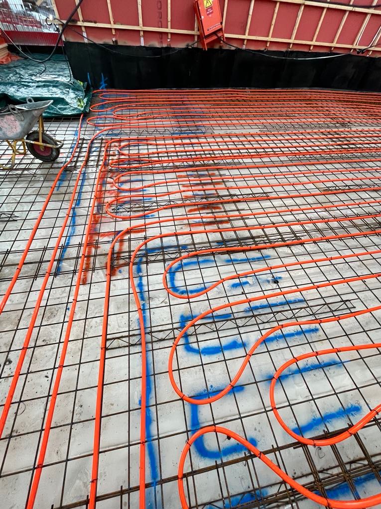 Pomarańczowe rurki instalacji podłogowej ułożone na siatce zbrojeniowej, z niebieskimi oznaczeniami na podłożu i taczką w tle.