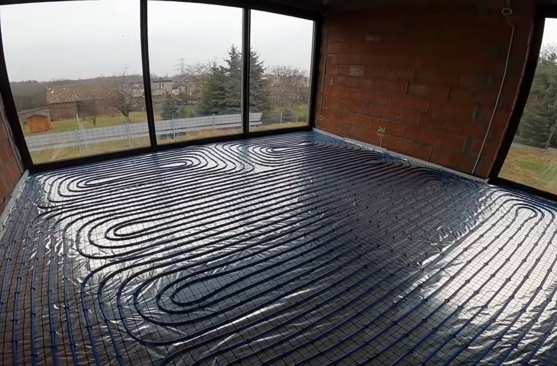 Niebieskie rury na srebrnej folii odblaskowej ułożone w pętle na podłodze w surowym stanie, widok z góry na instalację w pomieszczeniu z dużymi oknami.