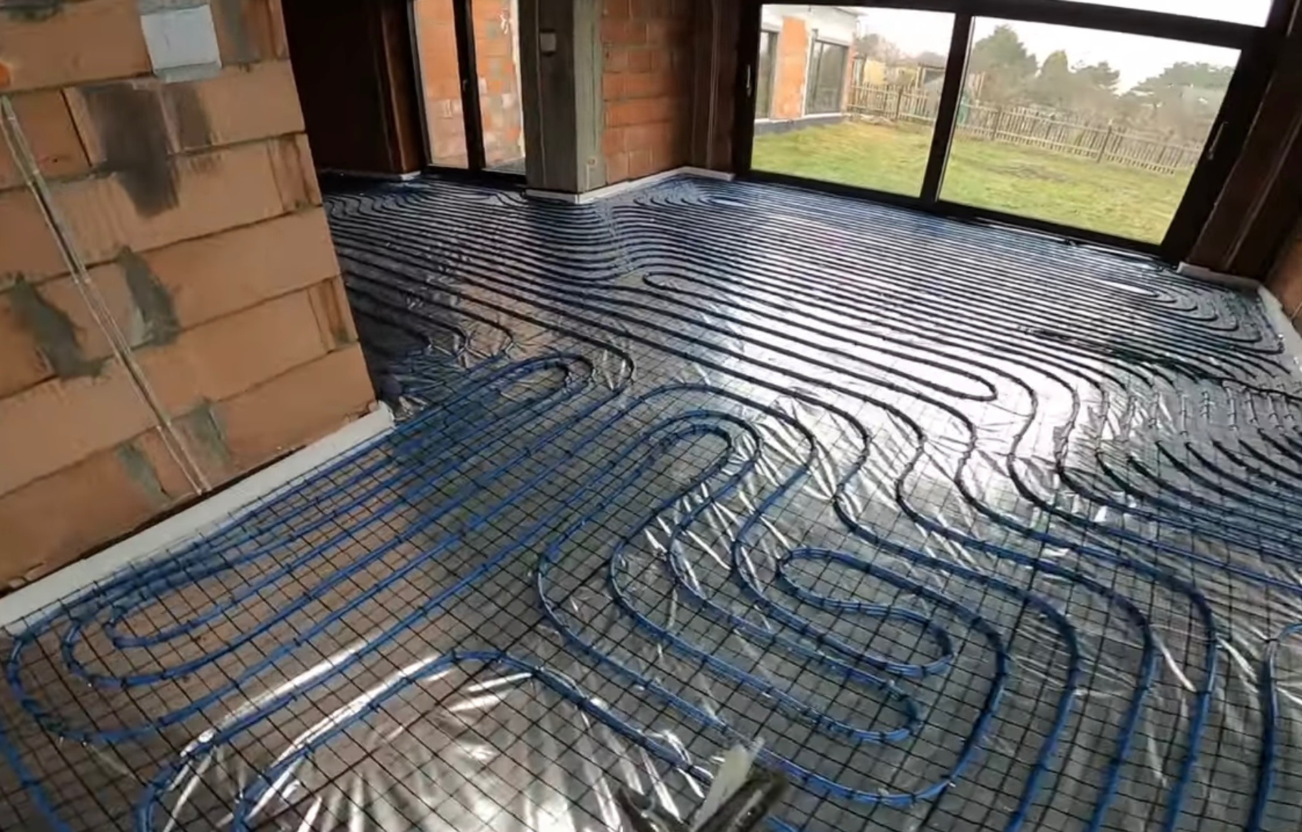 Niebieskie rurki układane w pętlach na srebrnej folii, tworzące system ogrzewania podłogowego w surowym wnętrzu z ceglanymi ścianami i dużymi oknami.