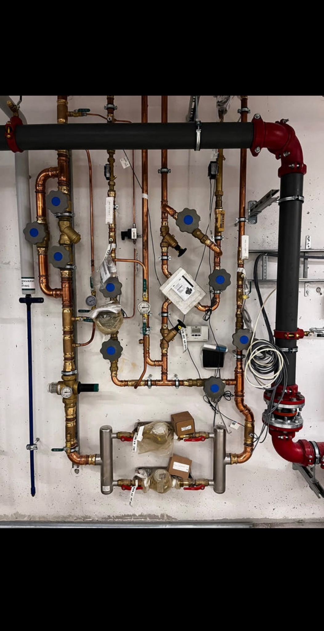 Rozbudowana instalacja z miedzianych rur, zaworów z niebieskimi pokrętłami i czarnych rur izolowanych, zamontowana na szarej, surowej ścianie. Czerwone elementy łączące rury w prawym górnym rogu.