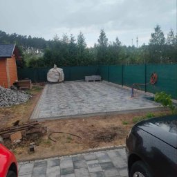 Usługi brukarskie - Ułożona kostka brukowa w odcieniach szarości na posesji z zielonym ogrodzeniem. Widoczny stóg materiałów budowlanych i samochody po bokach.