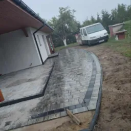 Ułożona kostka brukowa w odcieniach szarości z czarnym obrzeżem, tworząca ścieżkę prowadzącą do domu. W tle biały samochód dostawczy i zieleń.