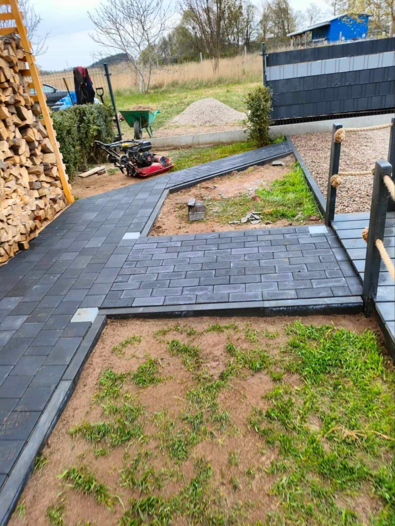 Ciemnoszara kostka brukowa ułożona w ogrodzie, widoczna ubijarka, hałda piasku i drewno na opał. Częściowo wykończony chodnik z kostki.