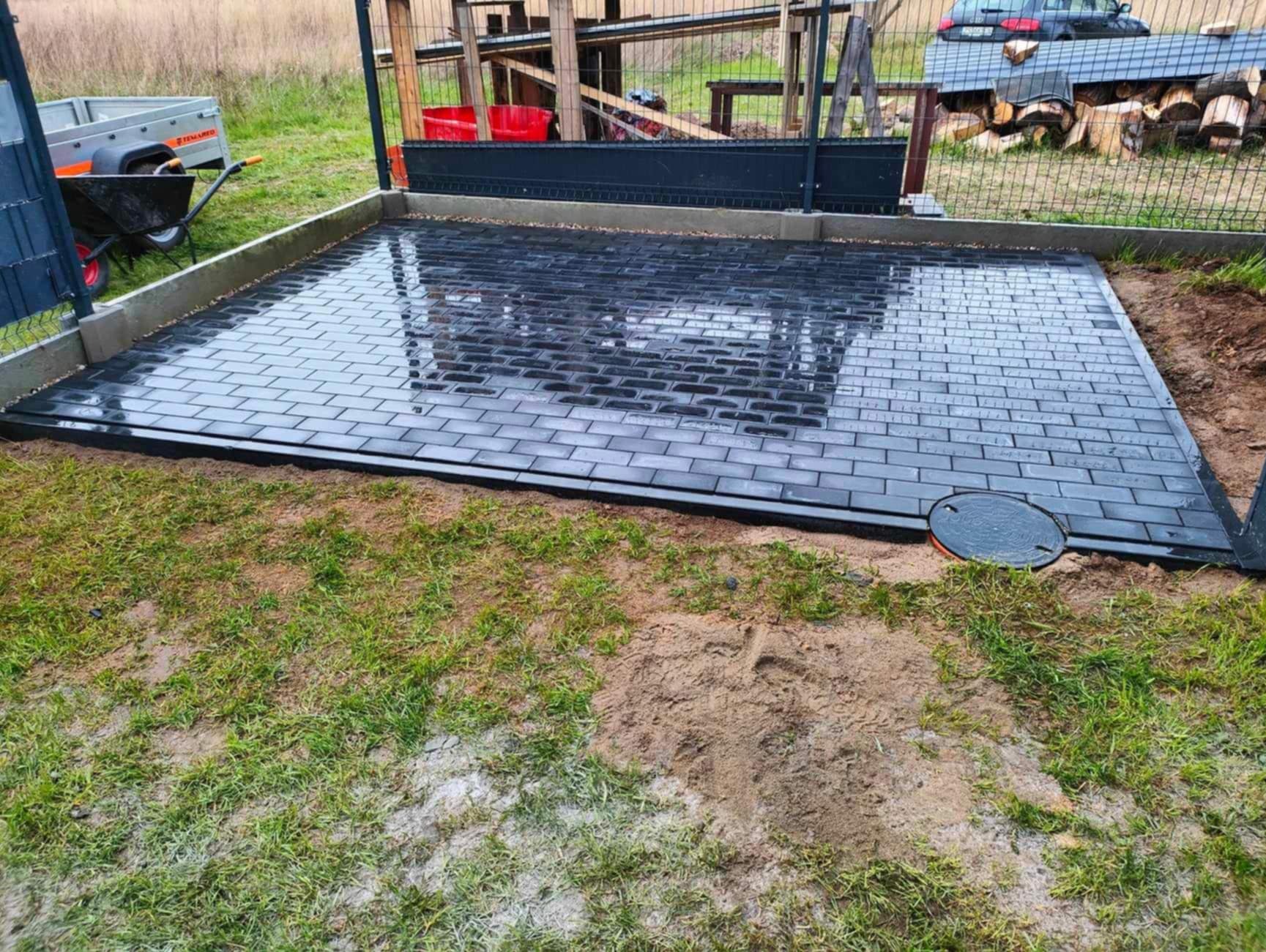 Świeżo ułożona, czarna kostka brukowa na posesji, z widocznym włazem kanalizacyjnym i fragmentem przyczepy. Kostka ułożona w klasyczny wzór cegiełki.