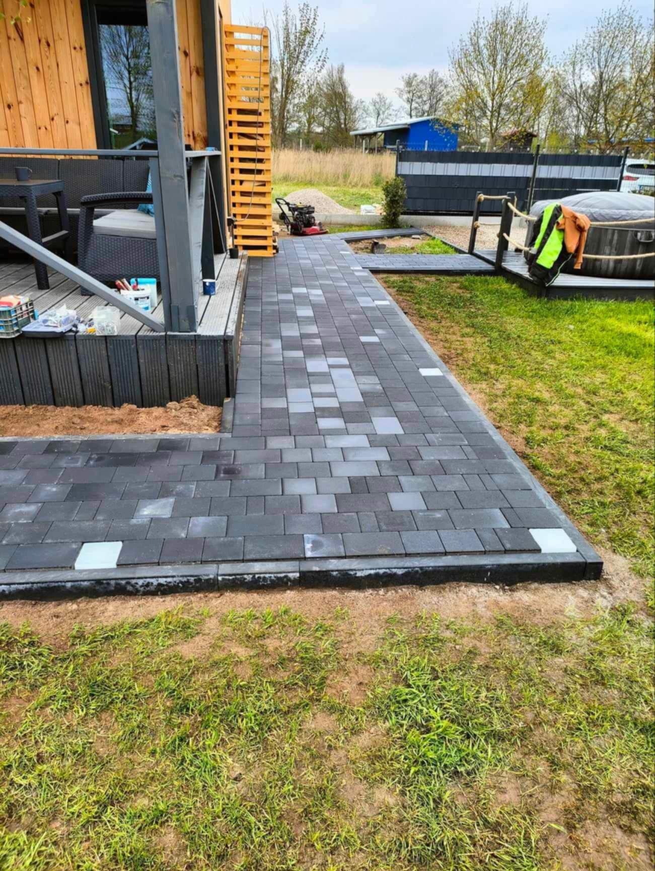 Ścieżka z kostki brukowej w odcieniach szarości, prowadząca od tarasu do jacuzzi na trawie, z widoczną zagęszczarką i elementami drewnianej architektury.