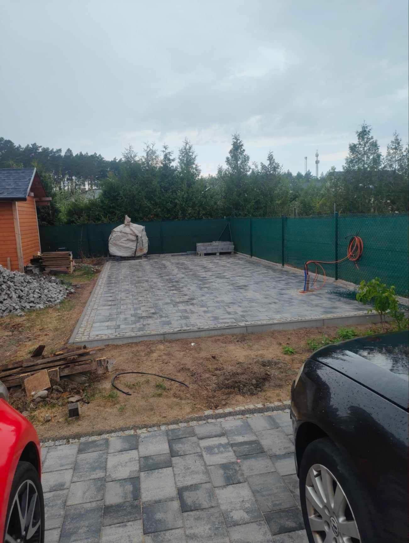 Ułożona kostka brukowa w odcieniach szarości na posesji z zielonym ogrodzeniem. Widoczny stóg materiałów budowlanych i samochody po bokach.