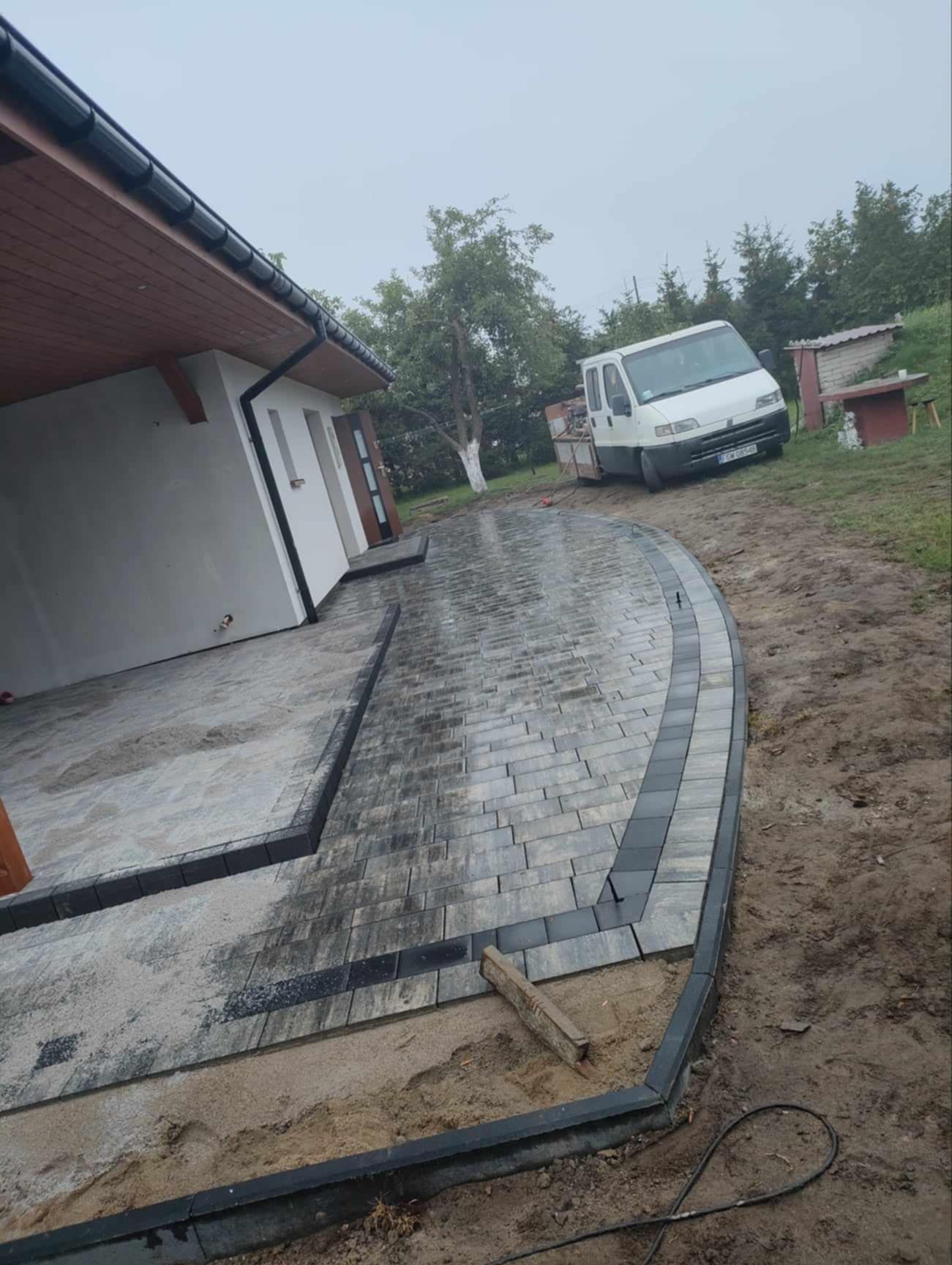 Ułożona kostka brukowa w odcieniach szarości z czarnym obrzeżem, tworząca ścieżkę prowadzącą do domu. W tle biały samochód dostawczy i zieleń.