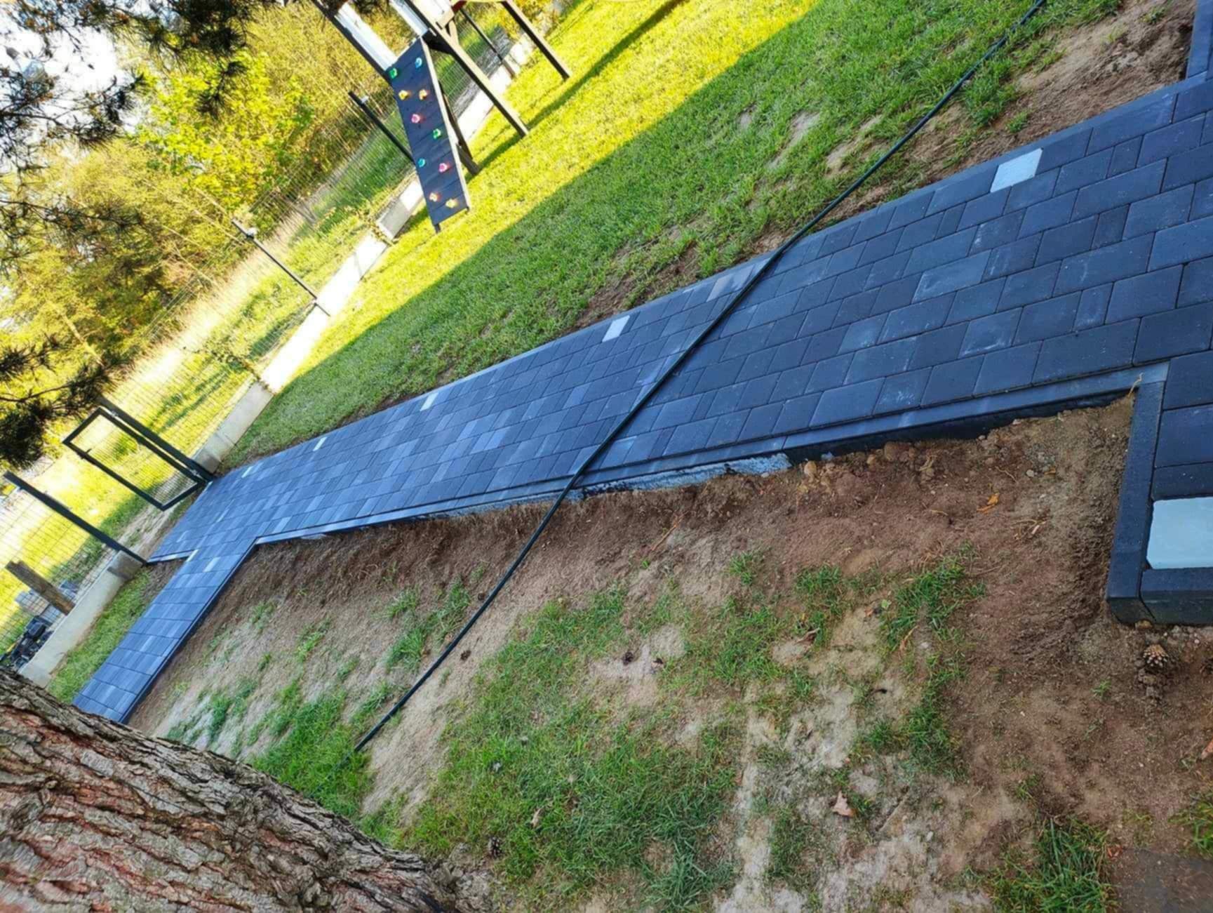 Ścieżka z ciemnej kostki brukowej w ogrodzie, widok z boku, z fragmentem pnia drzewa w lewym dolnym rogu i placem zabaw w tle. Kostka ułożona na podsypce.