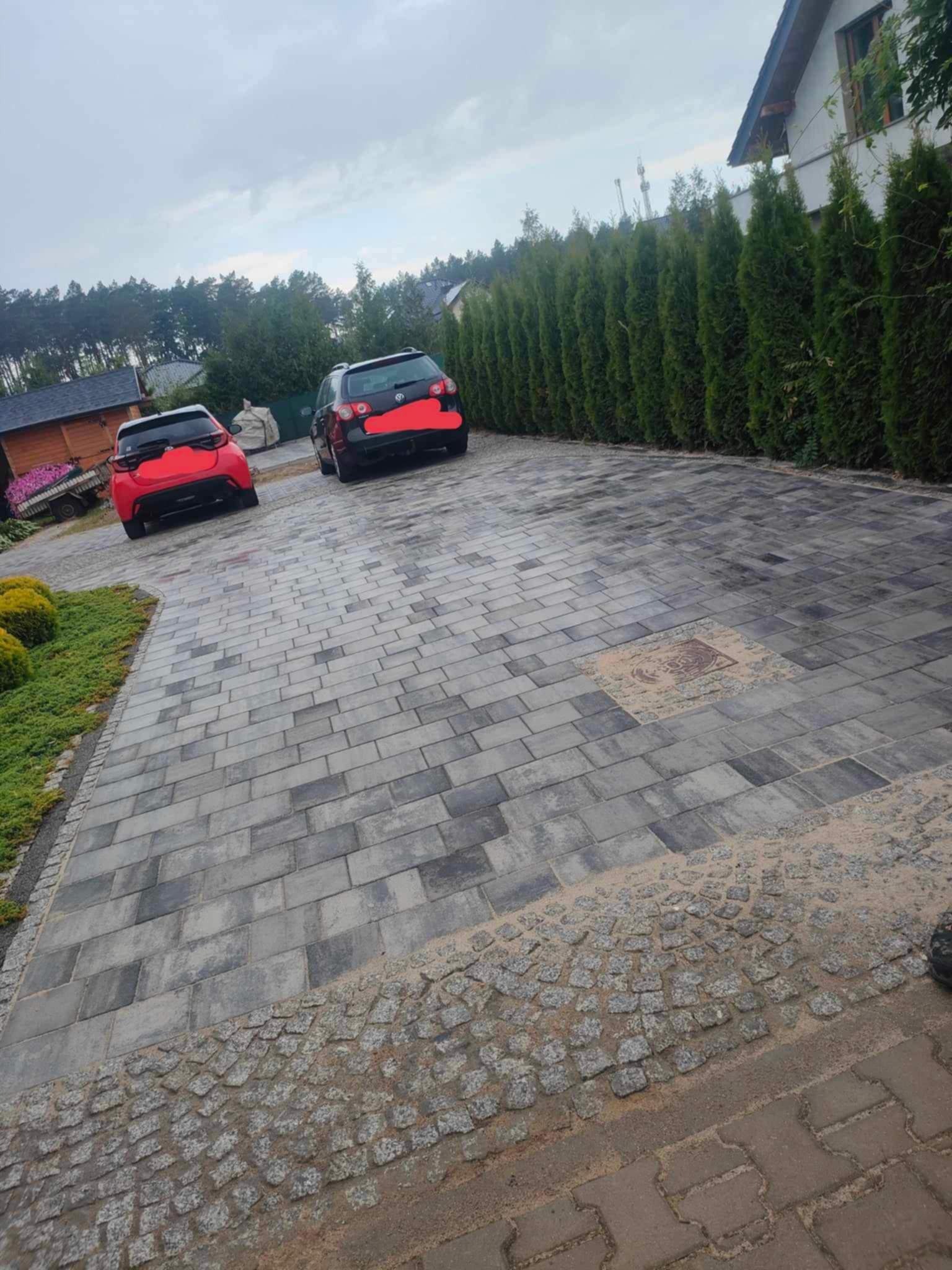 Ułożona kostka brukowa na podjeździe z dwoma samochodami w tle, obrzeża z kostki granitowej, widoczne studzienki rewizyjne i tuje wzdłuż ogrodzenia.
