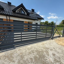 FOXsteel - Nowoczesna, przesuwna brama wjazdowa z poziomymi panelami, zamontowana przy posesji z czarnym dachem i panelami słonecznymi, częściowo otwarta, widoczny samochód.