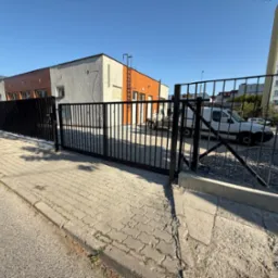 Czarna, przesuwna brama wjazdowa z metalowymi prętami na tle budynków i zaparkowanego białego samochodu dostawczego. Widok z zewnątrz posesji.