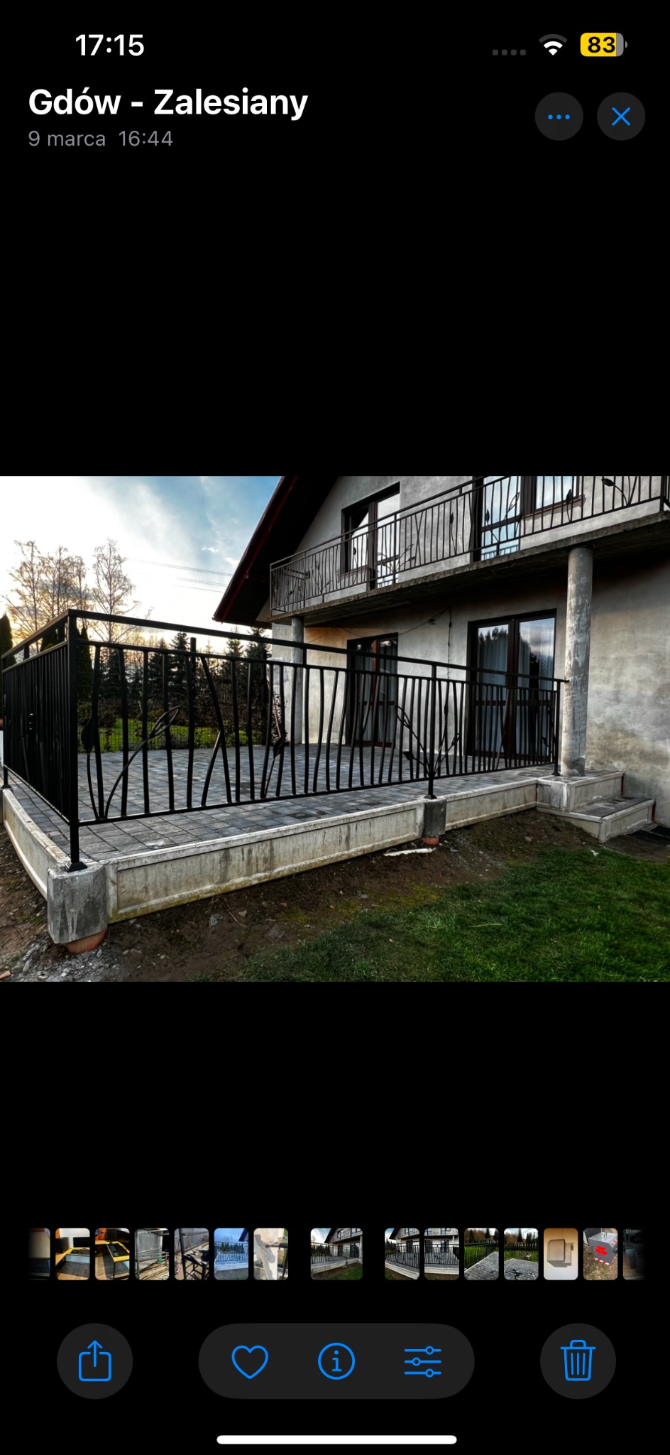 Czarna metalowa balustrada z motywem liści na tarasie domu jednorodzinnego, widoczna również balustrada na balkonie na piętrze, szara kostka brukowa i trawnik.