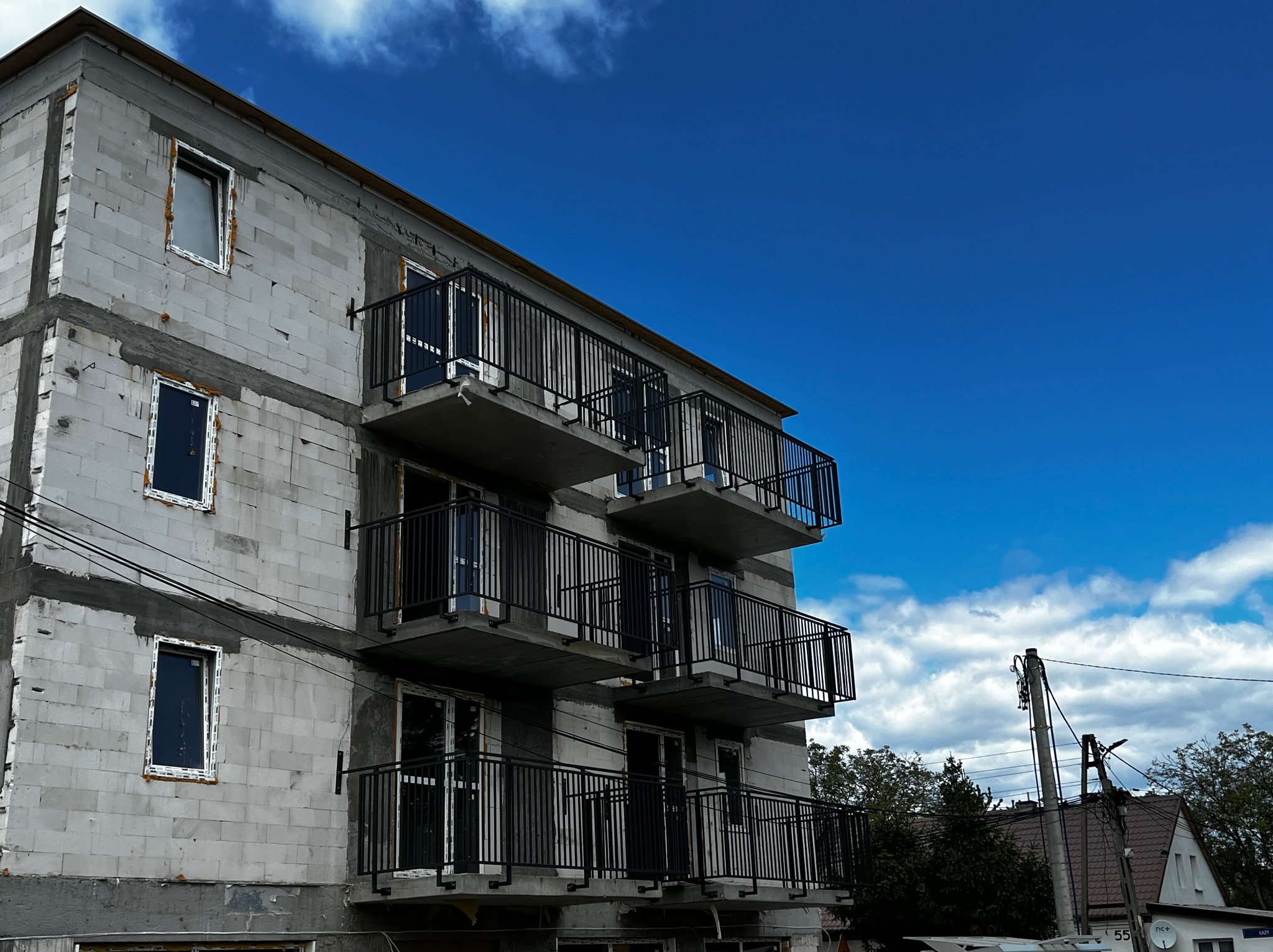 Surowe ściany budynku z zamontowanymi, czarnymi balustradami balkonowymi na tle błękitnego nieba z chmurami. Widok z dołu, podkreślający pionowość konstrukcji.