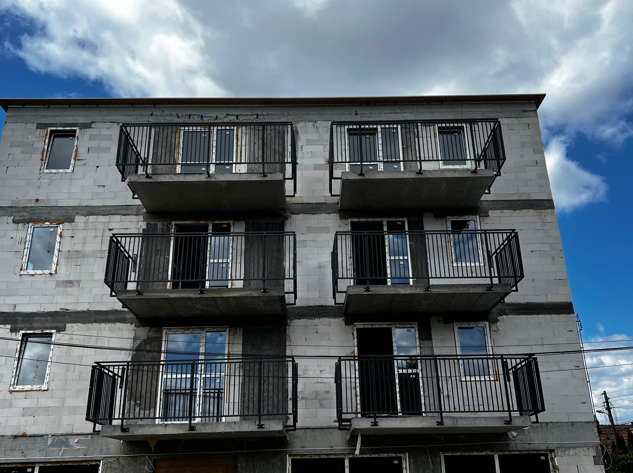 Wielopiętrowy budynek w stanie surowym z zamontowanymi czarnymi, metalowymi balustradami balkonowymi. Widoczne okna i fragment instalacji elektrycznej na tle nieba.