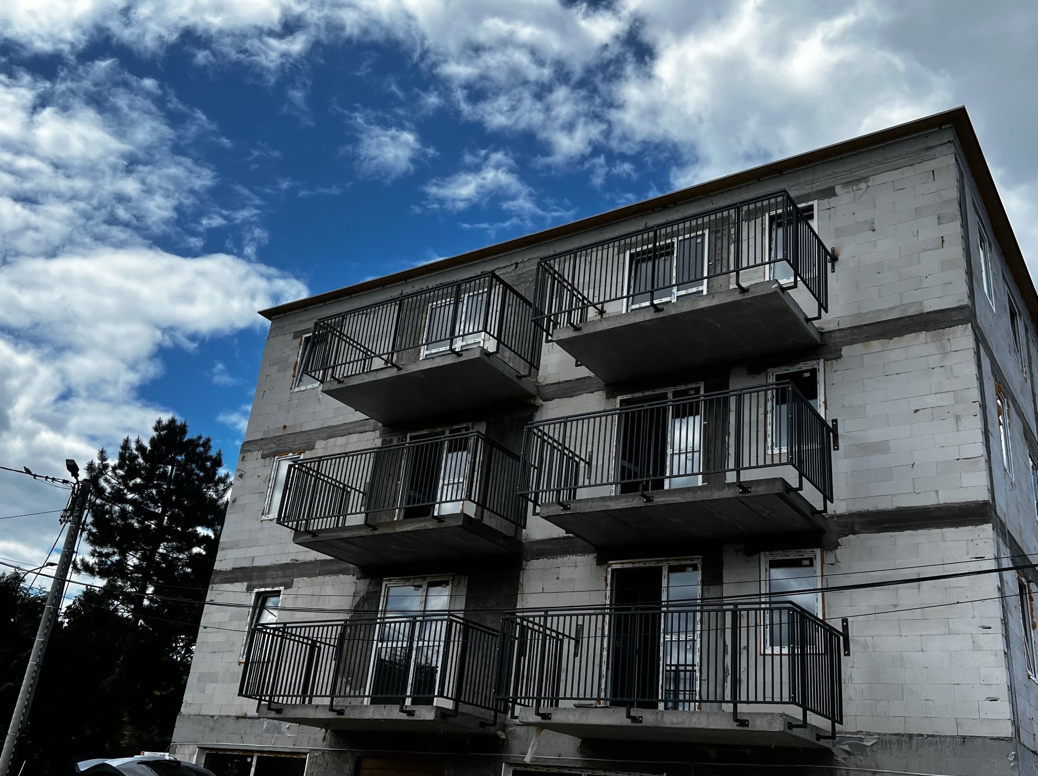 Surowe ściany budynku z zamontowanymi czarnymi, metalowymi balustradami balkonowymi, widziane z dołu na tle błękitnego nieba z chmurami.