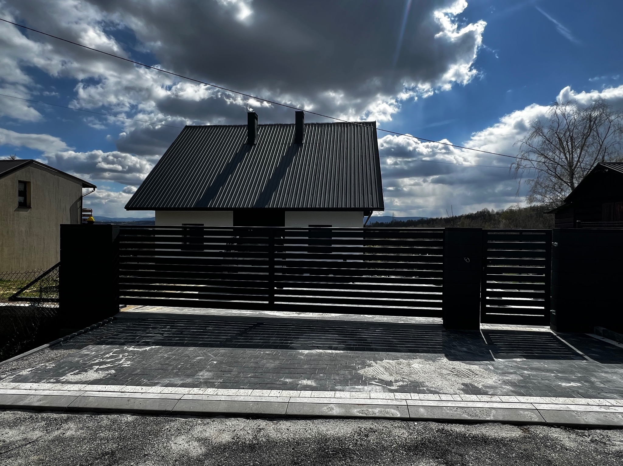 Nowoczesna, pozioma brama wjazdowa z czarnych profili na tle domu z grafitowym dachem i dekoracyjnej kostki brukowej. Słoneczny dzień, błękitne niebo z chmurami.