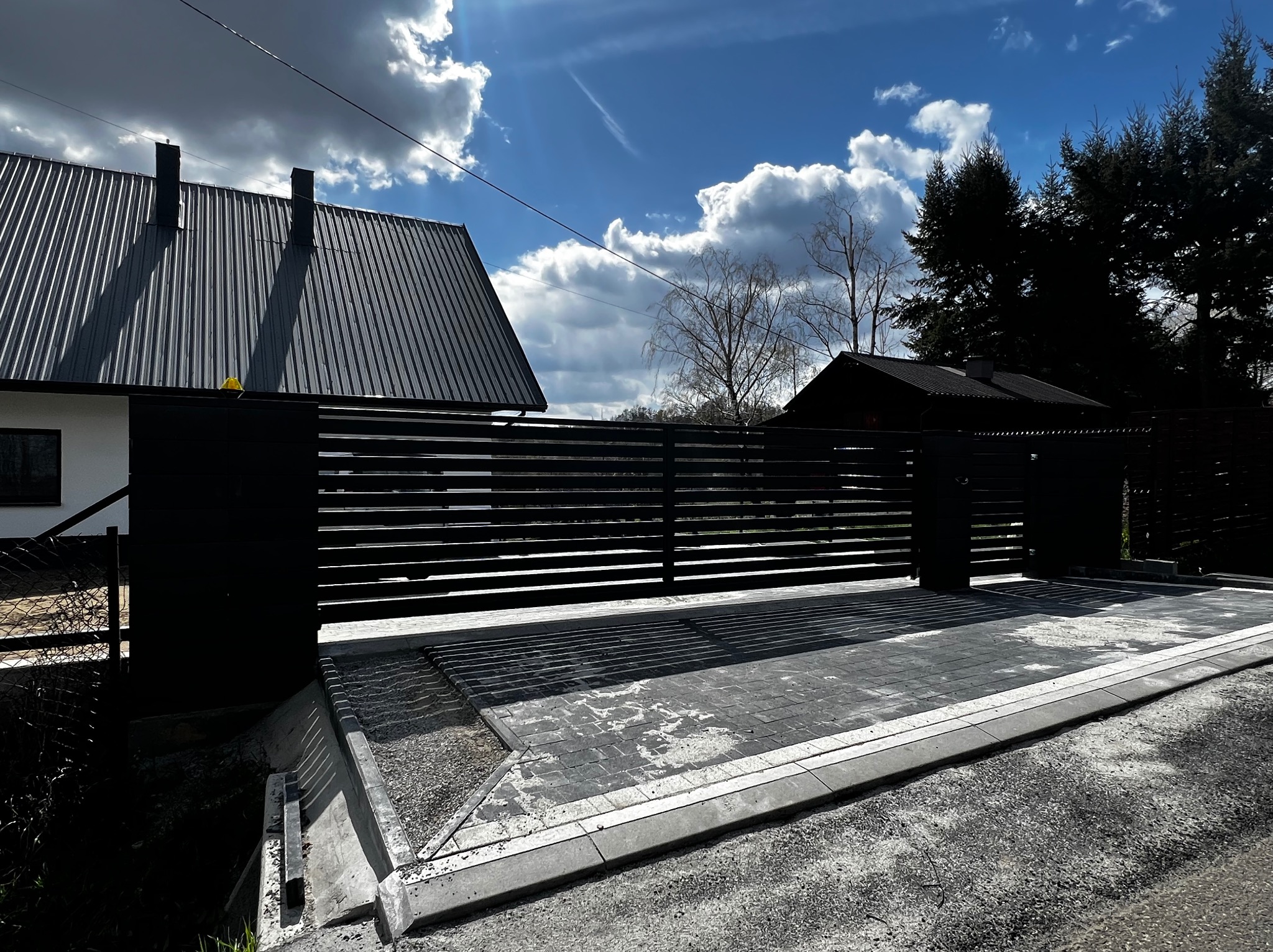 Nowoczesna, pozioma brama wjazdowa w kolorze czarnym, z automatyką, na tle domu z ciemnym dachem i błękitnego nieba z chmurami. Kostka brukowa przed bramą.