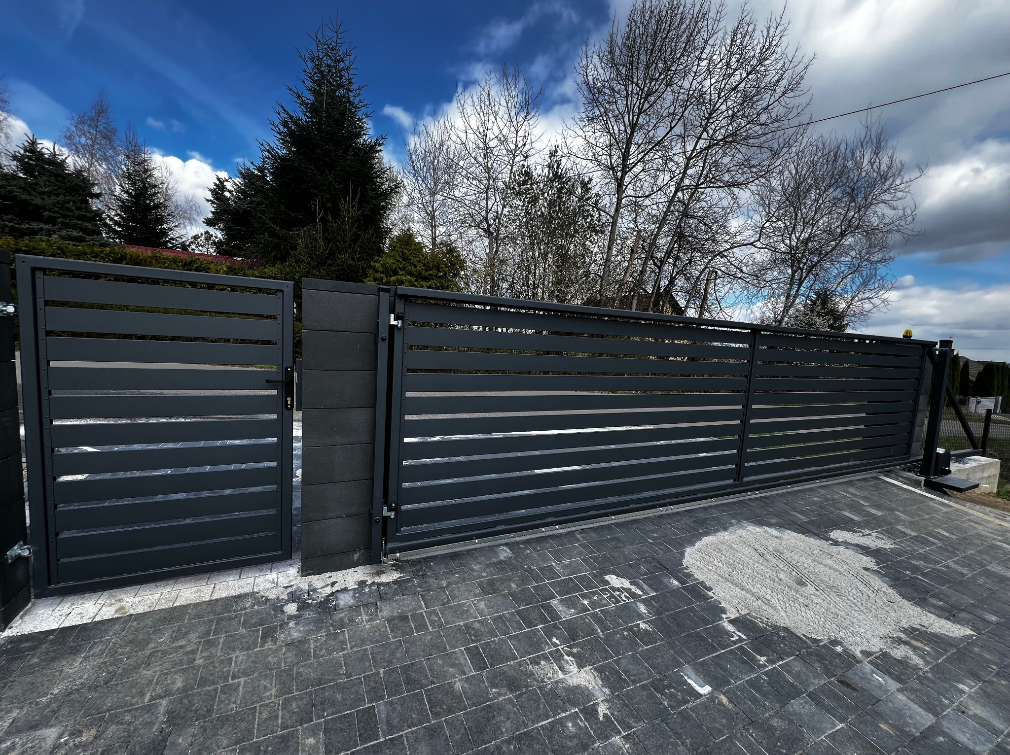 Nowoczesna, przesuwna brama wjazdowa w kolorze antracytowym z poziomymi panelami. Widoczna furtka i szara kostka brukowa na podjeździe, błękitne niebo w tle.