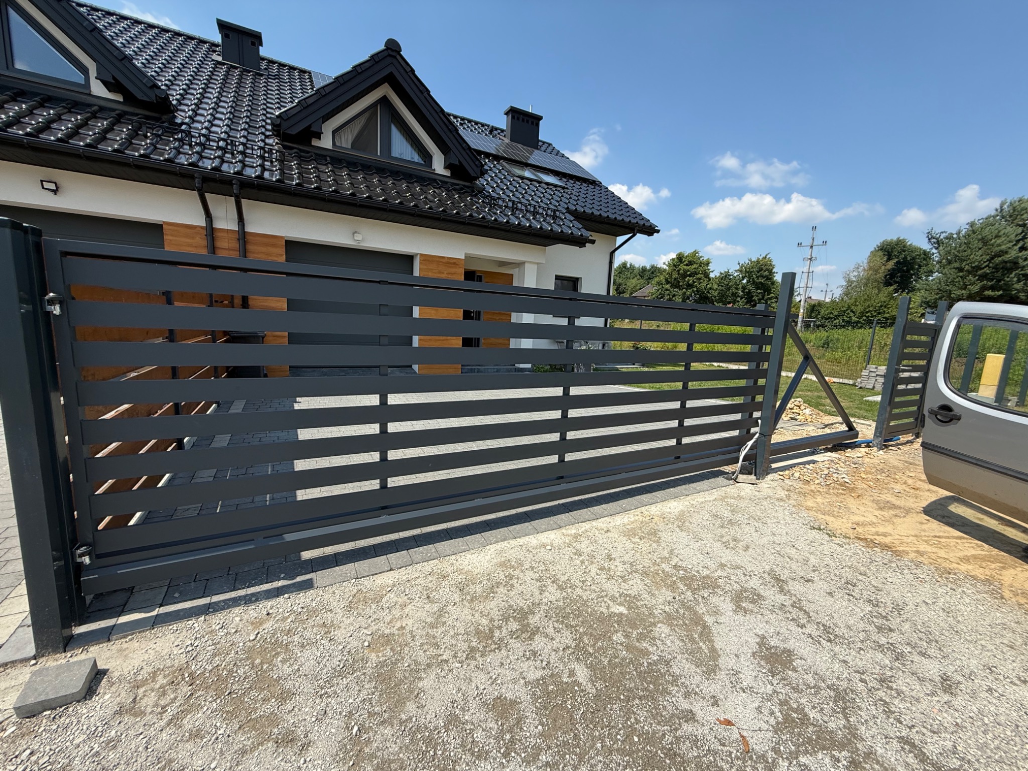 Nowoczesna, przesuwna brama wjazdowa z poziomymi panelami, zamontowana przy posesji z czarnym dachem i panelami słonecznymi, częściowo otwarta, widoczny samochód.
