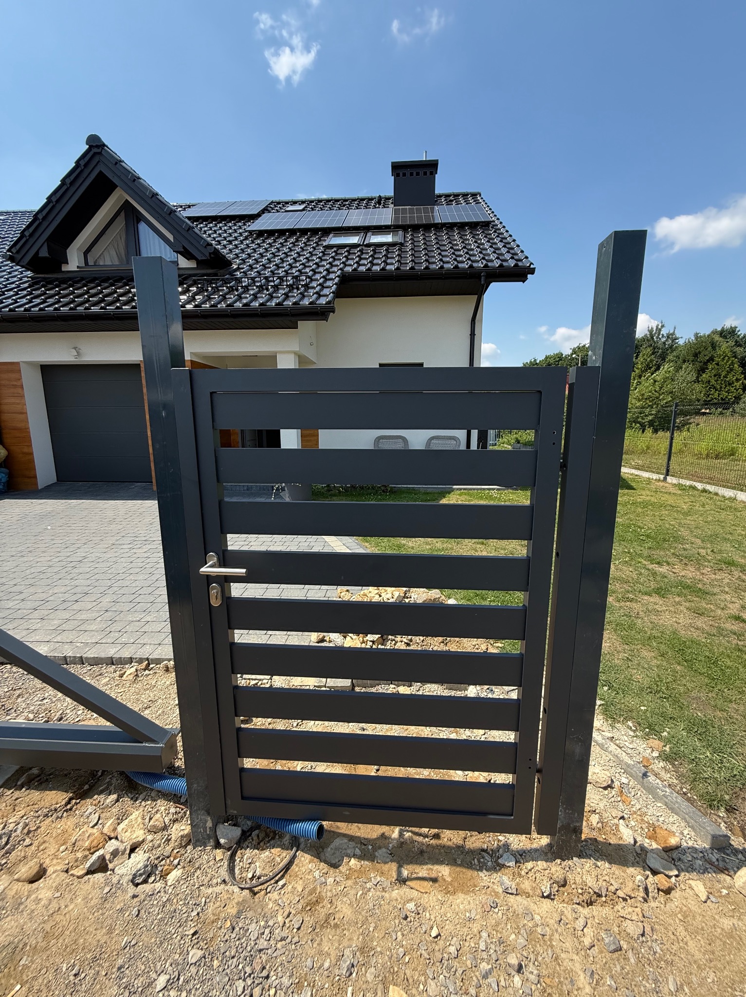 Nowoczesna, pozioma brama wjazdowa w kolorze antracytu w Zagórzanach. Widoczny dom z panelami słonecznymi i podjazd z kostki brukowej. Minimalistyczny design.