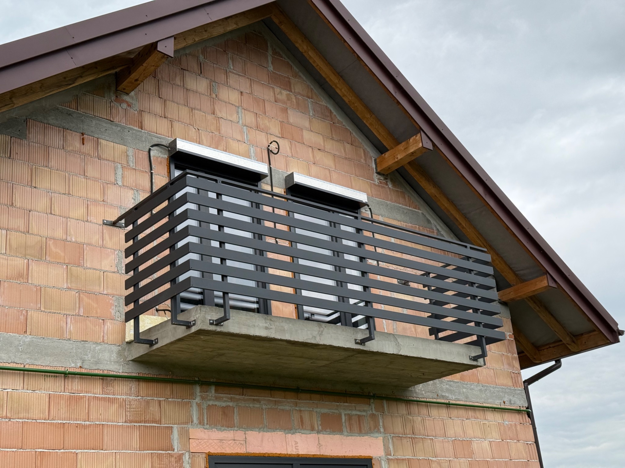 Nowoczesna, czarna balustrada balkonowa z poziomych profili, zamontowana na surowym, ceglanym budynku w trakcie budowy, z widocznym oknem i elementami dachu.