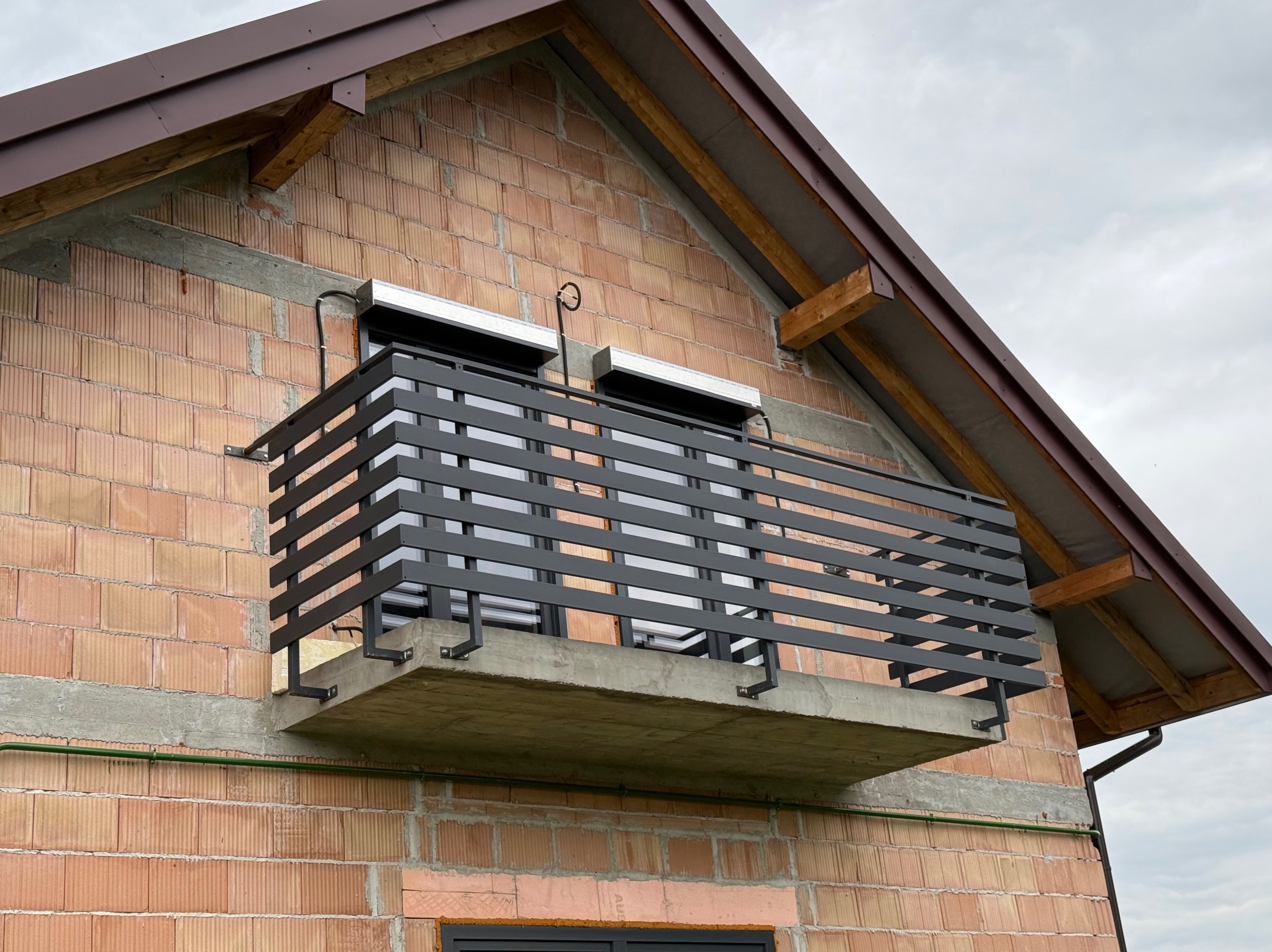 Nowoczesna, pozioma balustrada balkonowa z ciemnego metalu, zamontowana na betonowej płycie balkonu w surowym stanie, na tle ceglanego budynku w trakcie budowy.