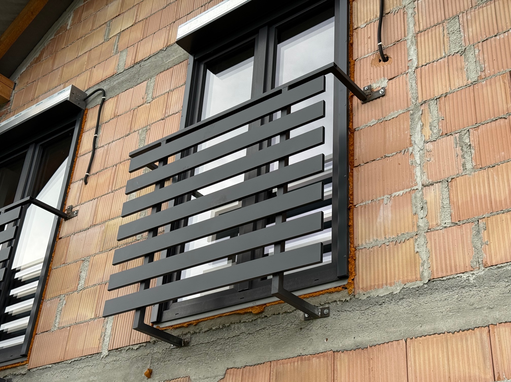 Nowoczesna, antracytowa balustrada okienna z poziomych profili, zamontowana na surowej, ceglanej ścianie budynku w trakcie budowy, widoczna z bliska.