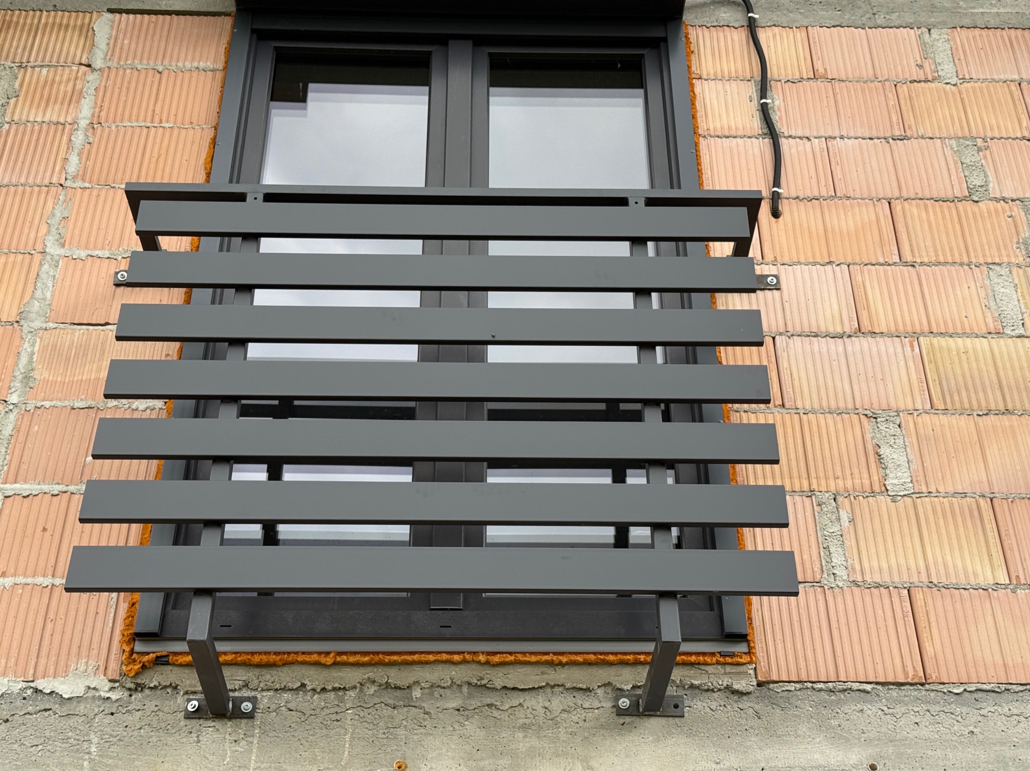 Szara, pozioma balustrada okienna z metalowych płaskowników, zamontowana na elewacji z cegły kratówki i betonu, widok z bliska.