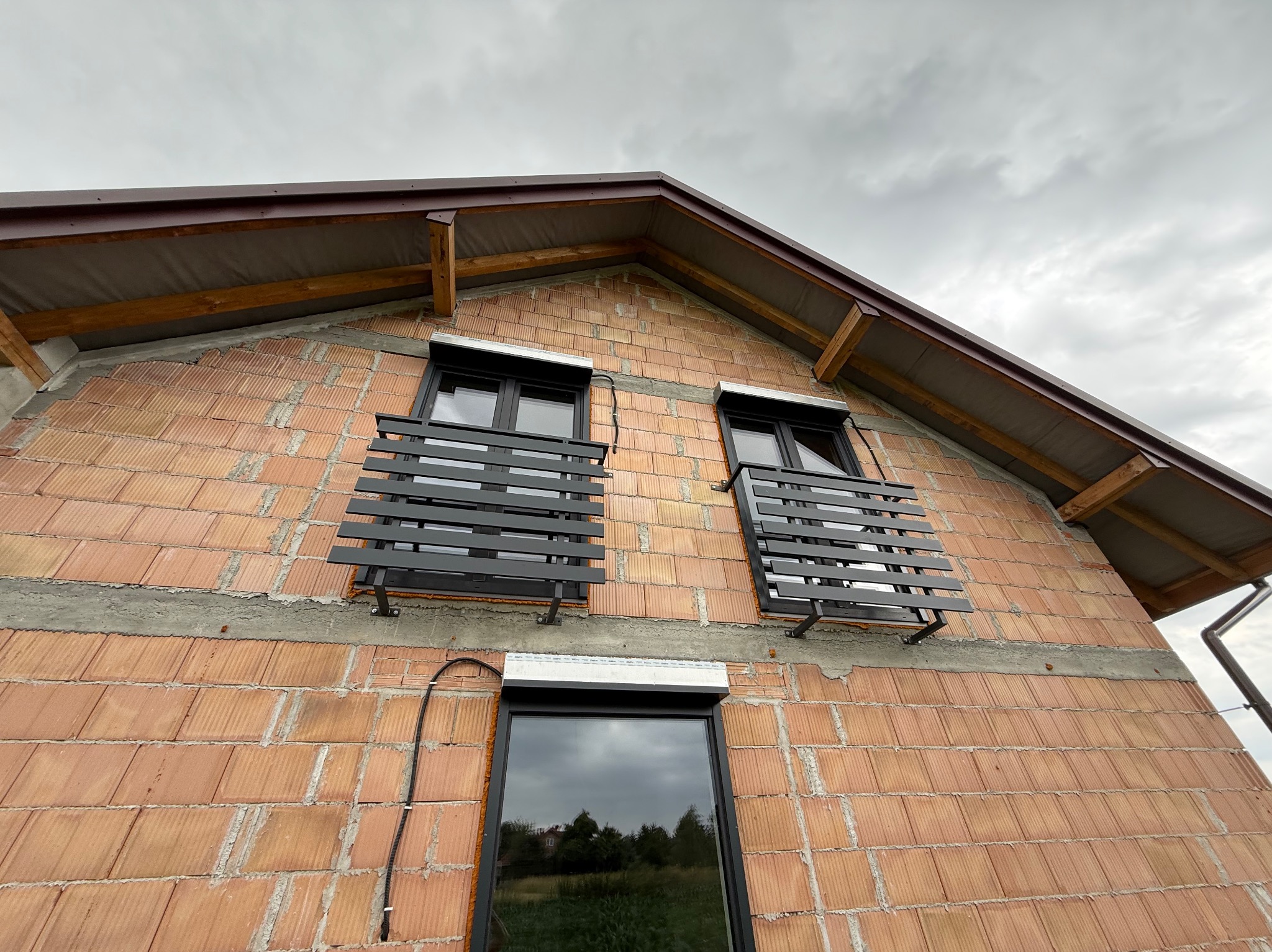 Surowy budynek z cegły z zamontowanymi czarnymi, metalowymi balustradami okiennymi i roletą. Widok z dołu na szary, pochmurny dzień.