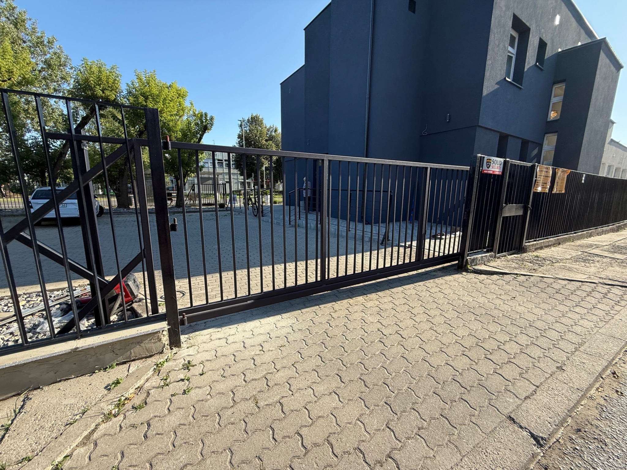 Zamknięta, przesuwna brama wjazdowa z metalu, z widocznym mechanizmem po lewej stronie. W tle szary budynek i drzewa. Kostka brukowa na pierwszym planie.