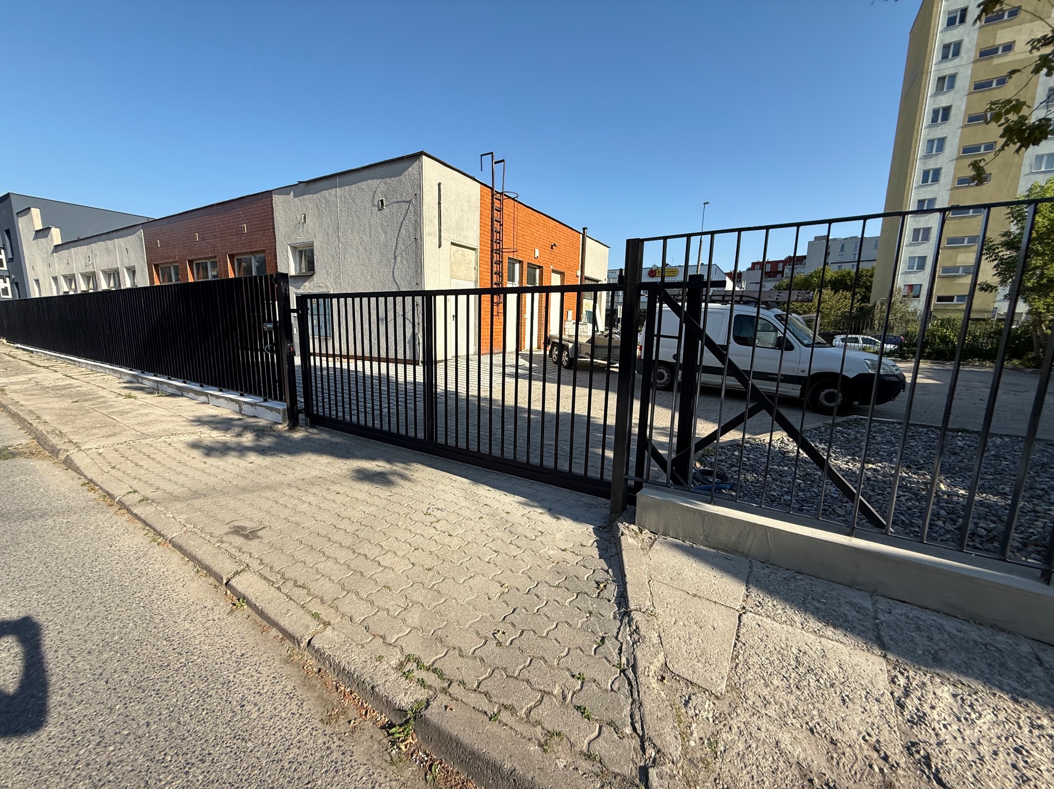 Czarna, przesuwna brama wjazdowa z metalowymi prętami na tle budynków i zaparkowanego białego samochodu dostawczego. Widok z zewnątrz posesji.