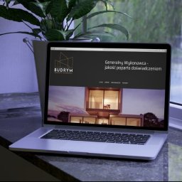 IYO CREATIVE SPÓŁKA Z OGRANICZONĄ ODPOWIEDZIALNOŚCIĄ - Laptop wyświetla stronę internetową firmy budowlanej 'BUDRYM' z wizualizacją nowoczesnego domu, ustawiony na parapecie obok doniczki z zieloną rośliną.